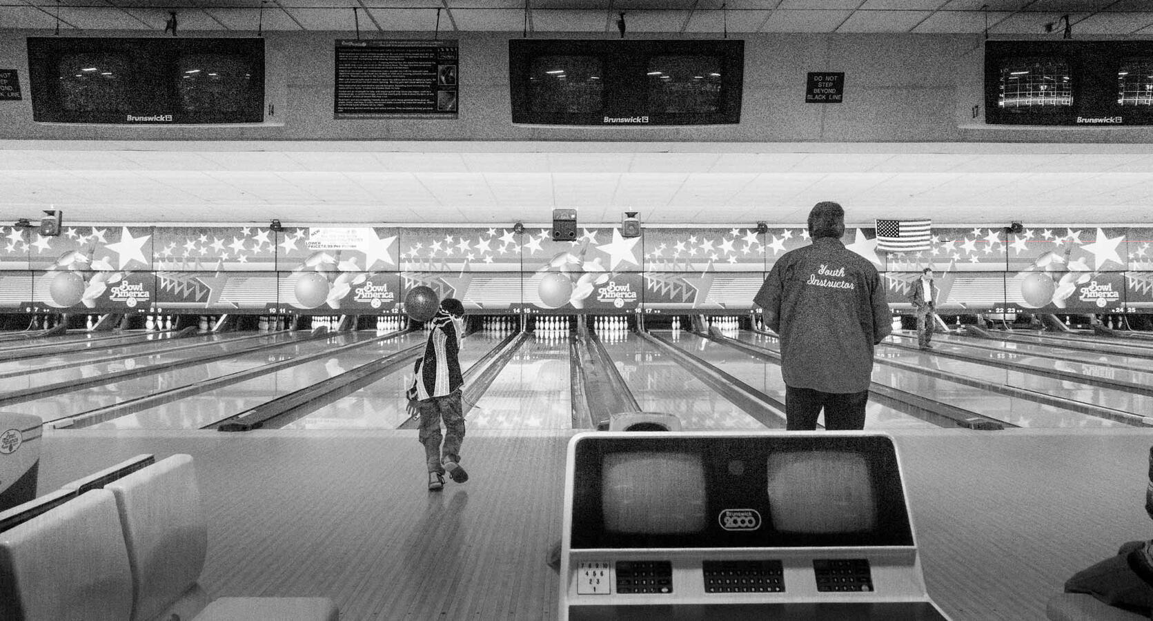 Black and white street photography from behind the bowling counter; a young boy is captured mid-motion as he releases a bowling ball down the lane under the watchful eye of a Youth Instructor.