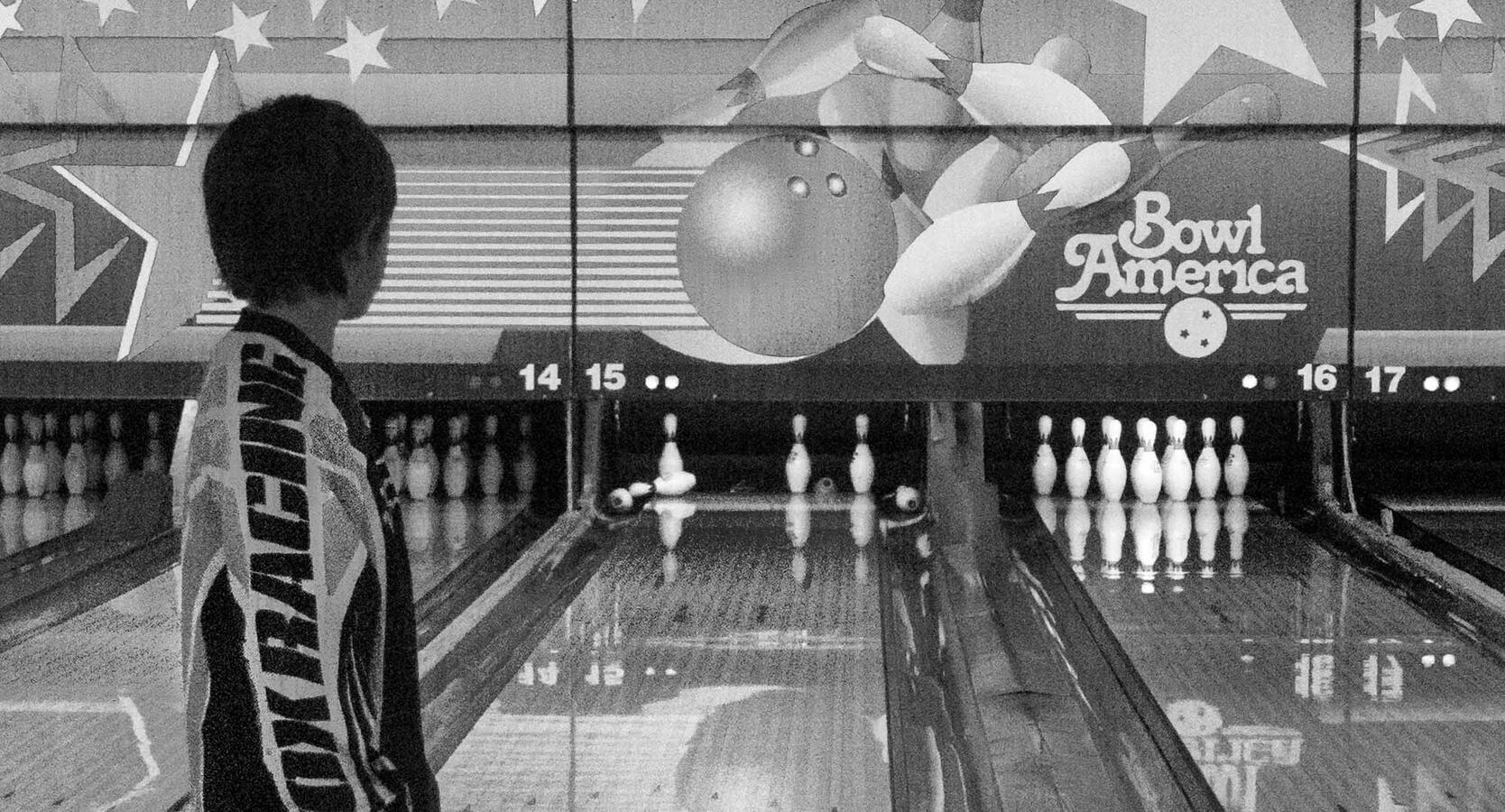 Black and white street photography showing the young boy standing at the end of the lane, looking down toward a difficult split of bowling pins remaining after his throw.
