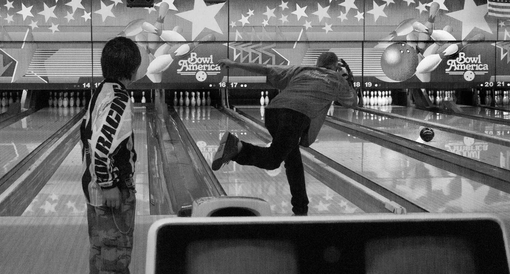 Black and white street photography where a bowling instructor is captured mid-release; due to the camera angle, his ball appears to be traveling down the wrong lane toward the pins on the right, while the young student watches from the left.