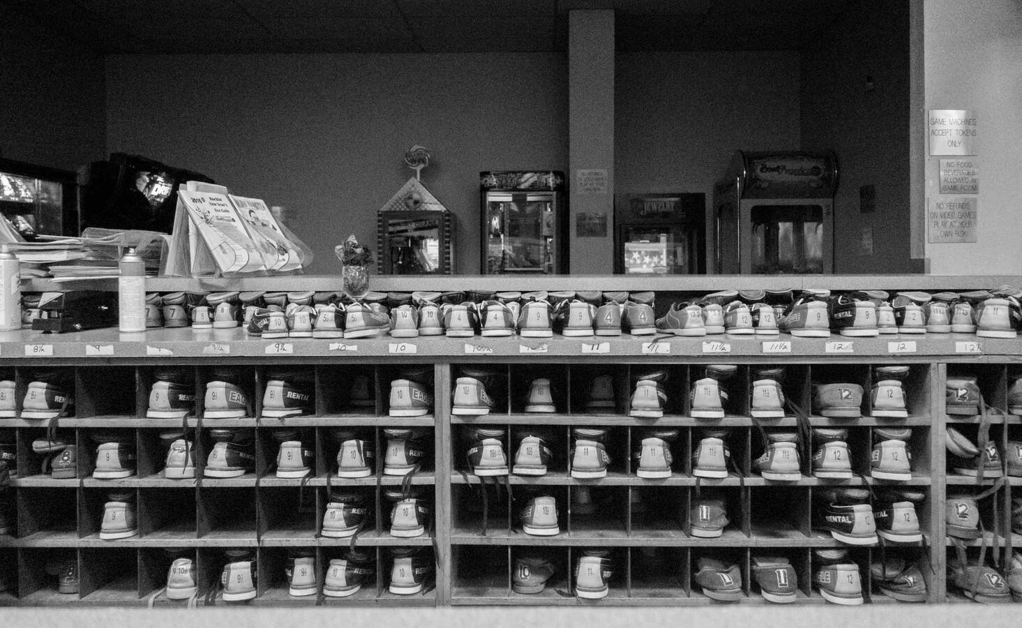 Black and white street photography of rental bowling shoes organized in wooden cubbies at a bowling alley service counter.