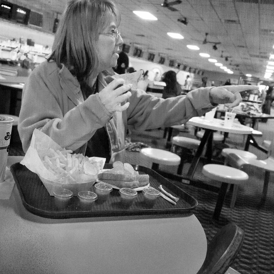 Black and white street photography of a woman at a bowling alley table eating a sandwich; she is turned away from the bowling lanes, pointing toward something off-camera in the opposite direction.