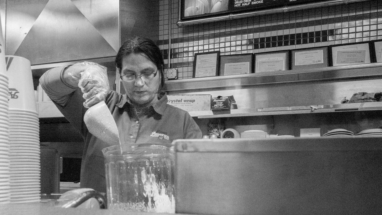 Black and white street photography from behind the counter of a bowling alley snack bar; a staff member wearing glasses and plastic gloves is focused on pouring ingredients into a large clear container.