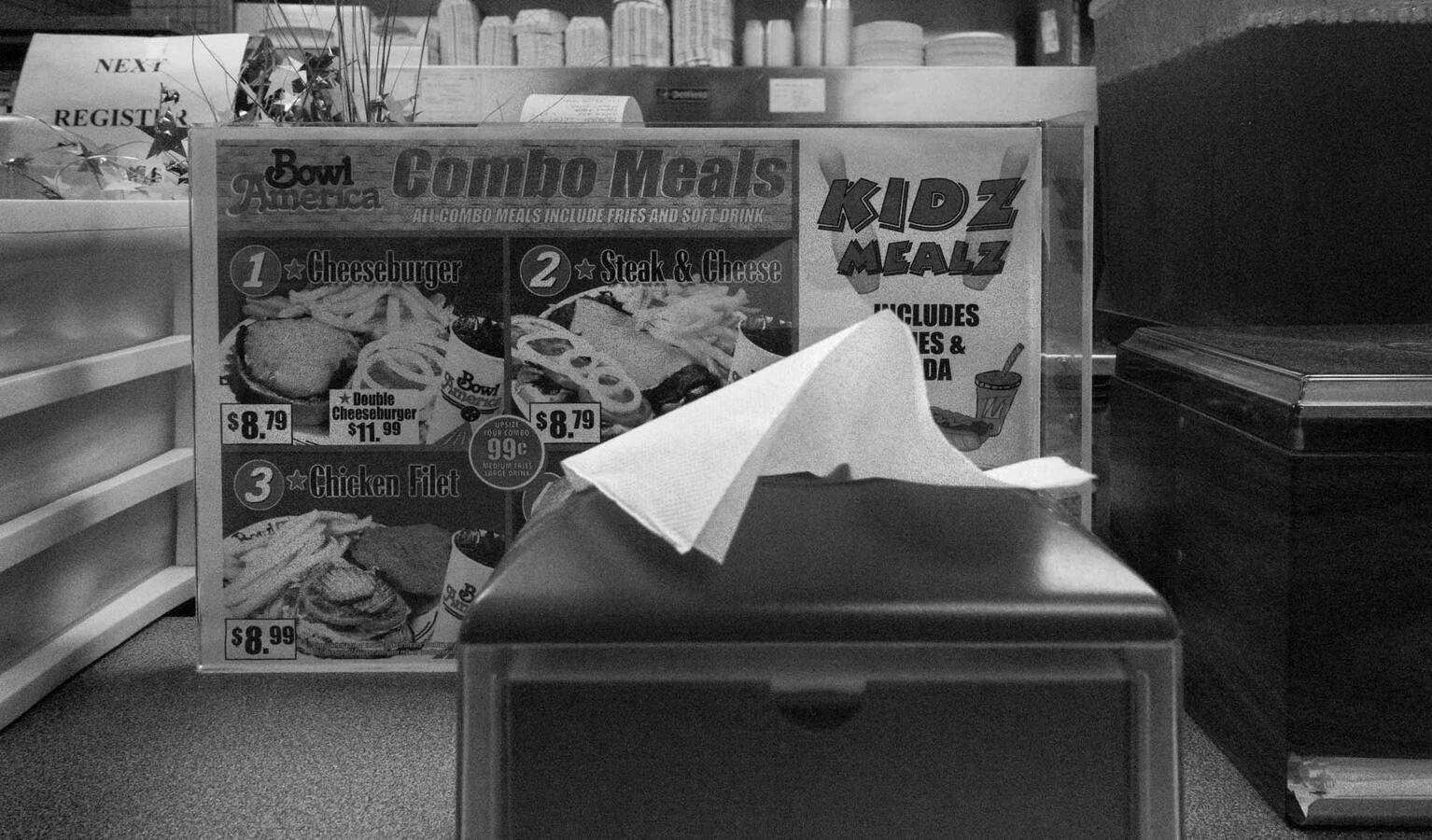 Close-up black and white street photography of a napkin dispenser in the foreground with a "Combo Meals" and "Kidz Mealz" menu sign in the background at a bowling alley.