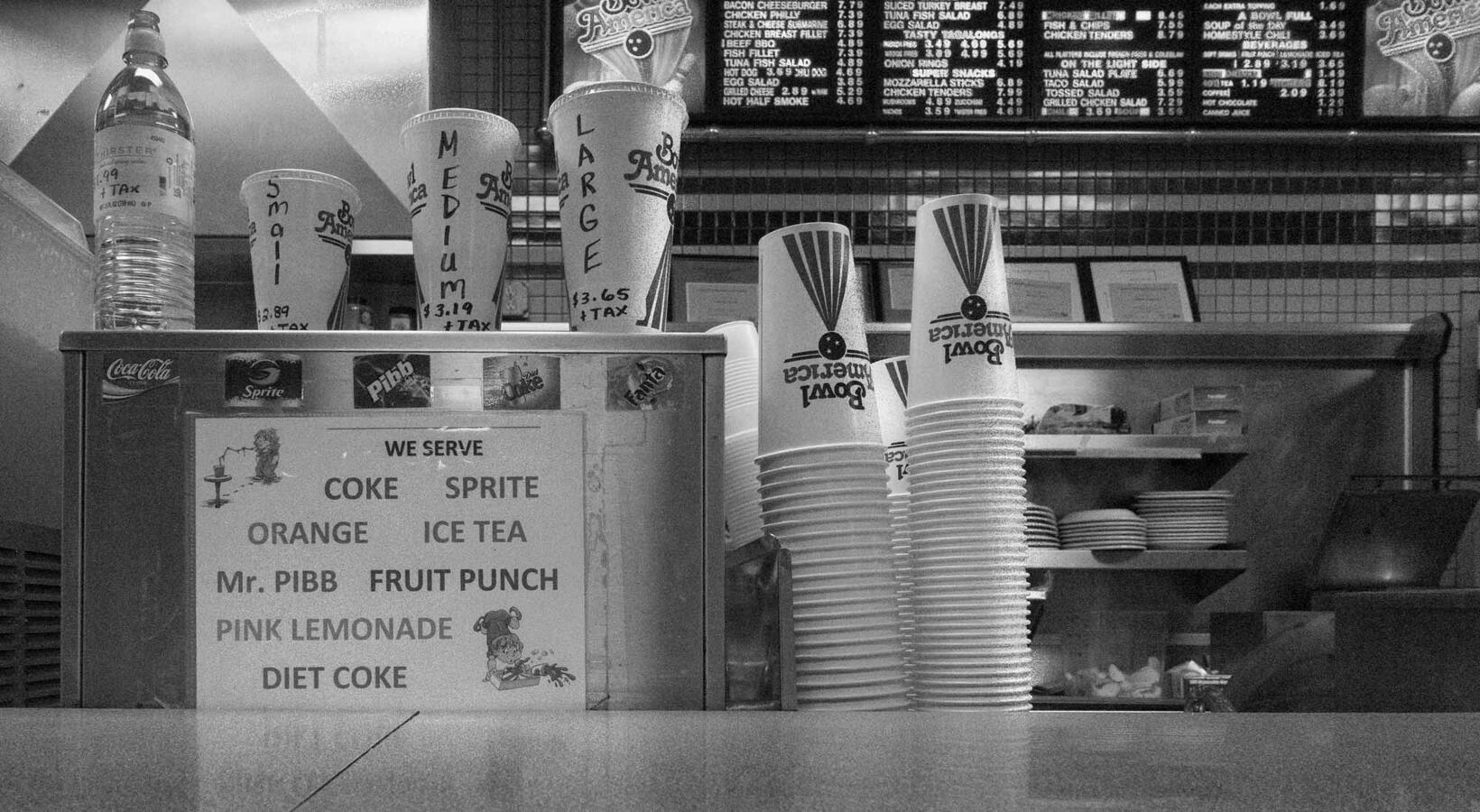 Black and white street photography of a bowling alley snack bar counter featuring stacked paper cups, a soda fountain menu, and hand-written pricing on drink displays.