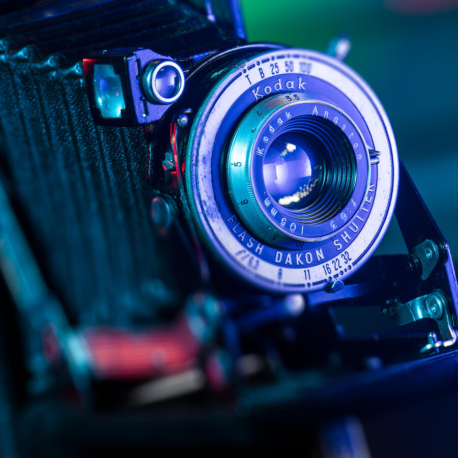 A close-up studio photograph of a vintage Kodak Bellows camera. Vibrant cyan and magenta light painting highlights the intricate details of the lens, shutter settings, and the accordion-style bellows, creating a modern, high-contrast aesthetic.