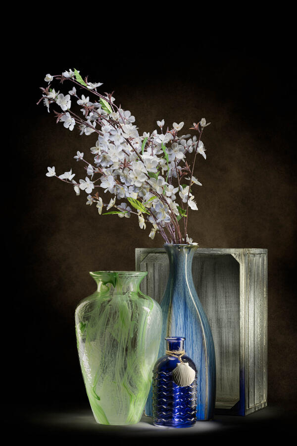 A vertical studio light painting of a vibrant green swirled glass vase, a tall blue textured vase, and a small dark blue bottle with a white seashell tied to its neck. Flowering white branches spill from the blue vase, and the arrangement is set against a