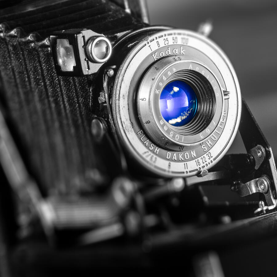 A dramatic black and white studio shot of a vintage Kodak Bellows camera. Selective coloring is used to highlight the camera's lens in a brilliant, luminous blue, drawing the eye to the glass while the textured metal and bellows remain in grayscale.