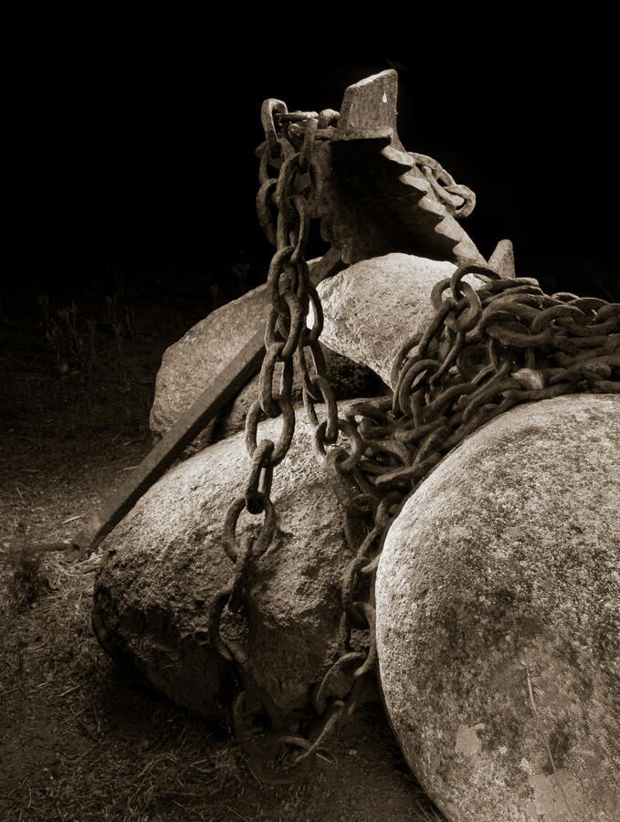 A vertical, close-up photograph of a rusted ship's anchor and heavy iron chains wrapped around large boulders on dry ground. Captured in sepia-toned black and white, the dramatic light painting emphasizes the pitted texture of the metal and the rough surfa