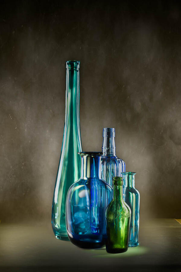 A studio light painting of five glass bottles in varying shades of cyan, deep blue, and emerald green. The lighting sculpts the sleek, elongated forms of the glassware against a smoky, atmospheric background.