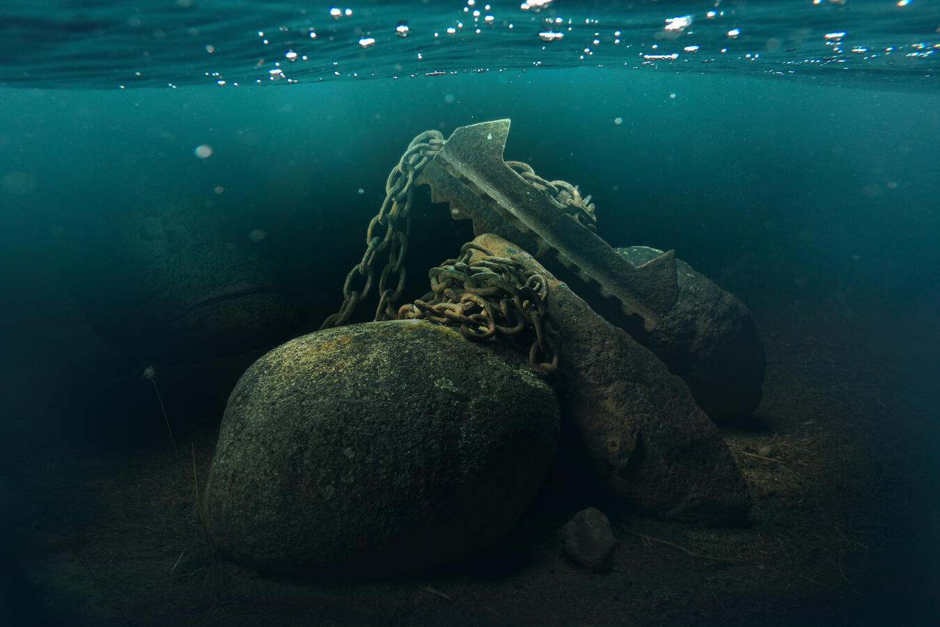 A surreal light-painted photograph of a massive, rusted ship's anchor chained to a boulder on a dry, earthy ground. Through a digital underwater overlay, a giant grouper fish is visible lurking in the deep shadows to the left, creating a dreamlike, submerg