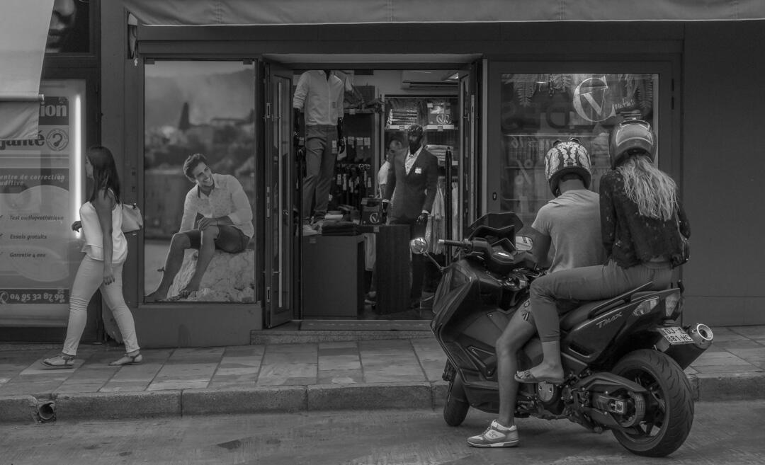 A complex black and white composition in Bastia. A photo of a model appears to look at a couple on a Vespa, while a mannequin inside a shop leans to look at a passing woman.