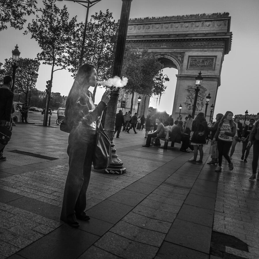 A cinematic black and white street photograph in Paris. A woman exhales a plume of smoke that catches the light, with the monumental Arc de Triomphe framed in the background.