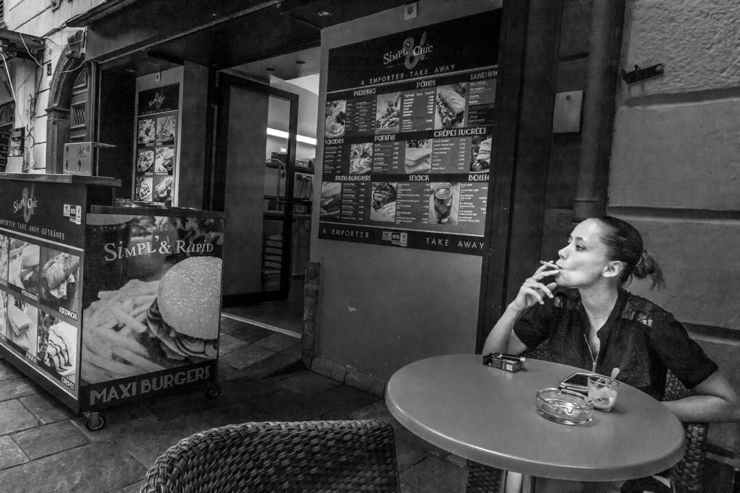 A candid black and white photograph of an employee taking a smoke break at a gelateria in Bastia, exhaling smoke next to a large "Simpl' & Rapid" advertisement.