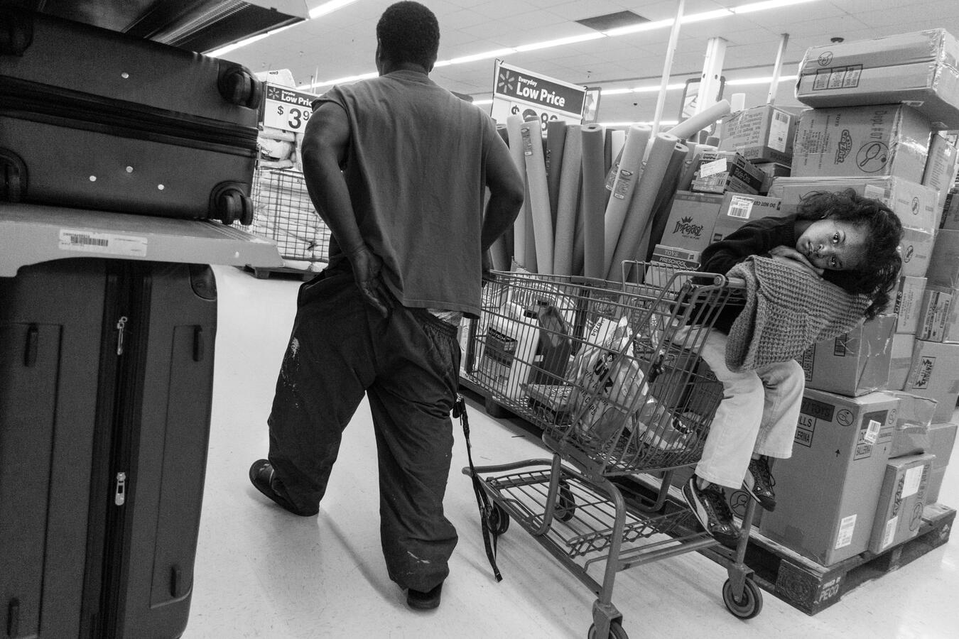 A humorous yet weary black and white scene in a Walmart. A tired young girl rests her head in a shopping cart as her father pulls the cart while adjusting his pants.