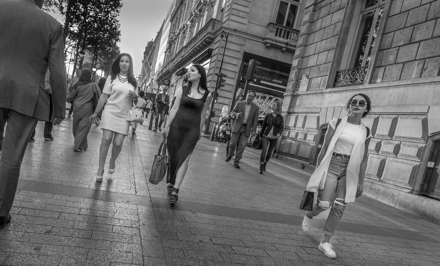 A black and white street scene on the Champs-Élysées. Well-dressed women walk past a woman in ripped jeans, highlighting the diverse fashion landscape of Paris.