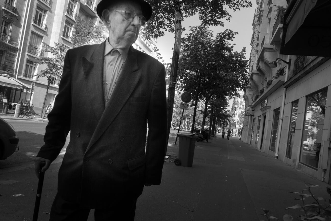 A dynamic black and white street photo in Paris. An elderly man in a suit and hat walks briskly with a cane, staring into a storefront window as the photographer passes him.