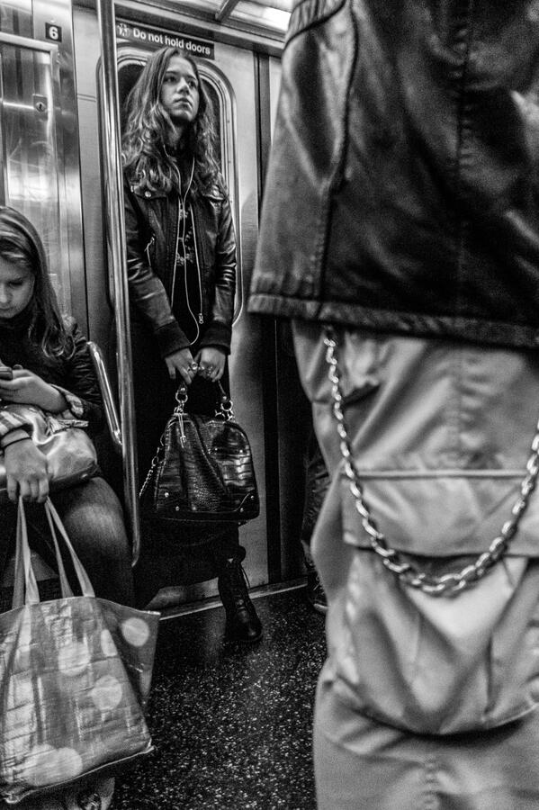 A vertical black and white street photograph inside a NYC subway car. A well-dressed woman in a leather jacket stands stoically, framed by the foreground texture of another commuter.