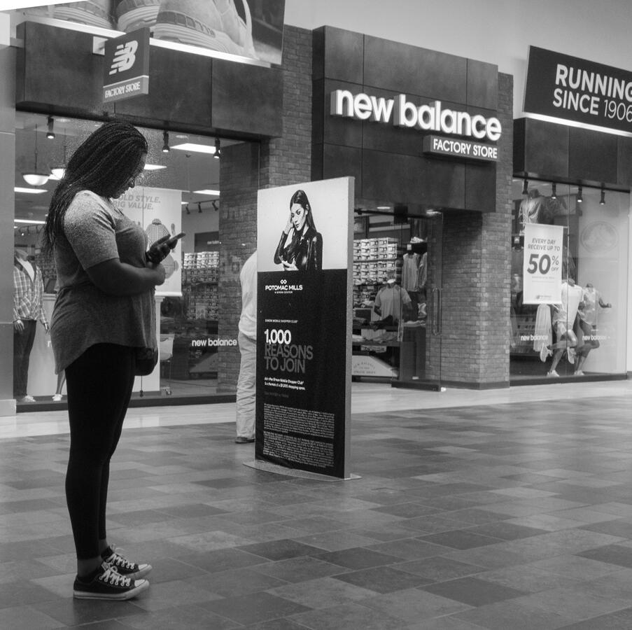A black and white street photo from a mall in Woodbridge. A woman stands looking at her phone directly in front of an advertisement featuring a woman in a similar pose.