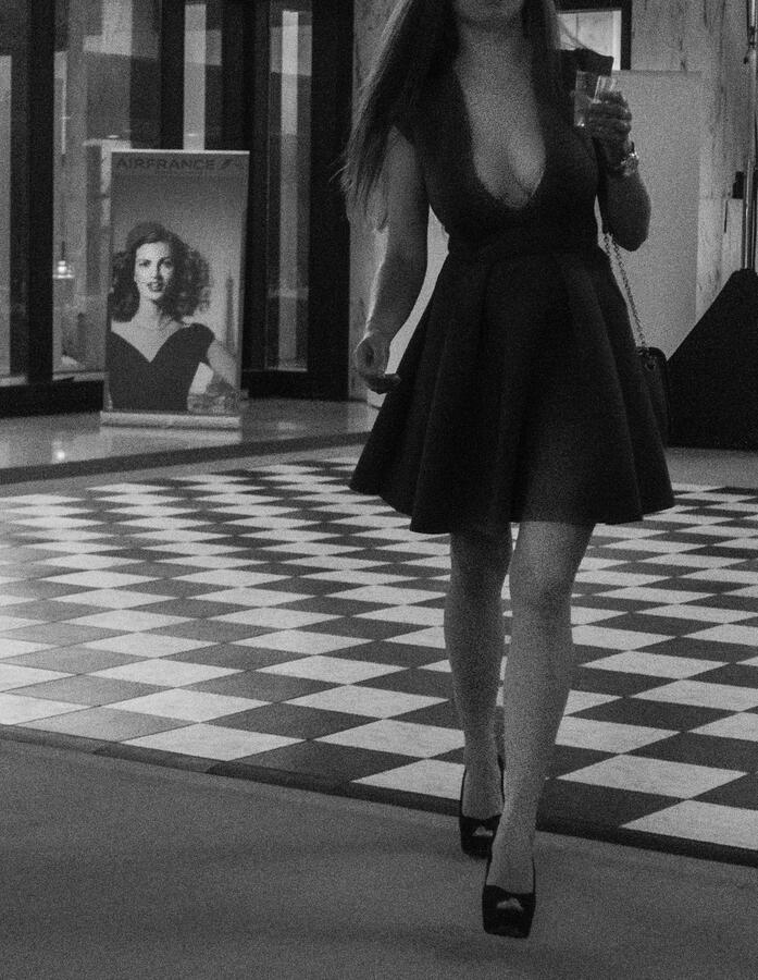 A high-contrast black and white street photograph at the French Embassy. A woman in a dark v-neck dress walks across a checkered floor, mirrored by an Air France poster in the background, completing the missing portions of the woman.