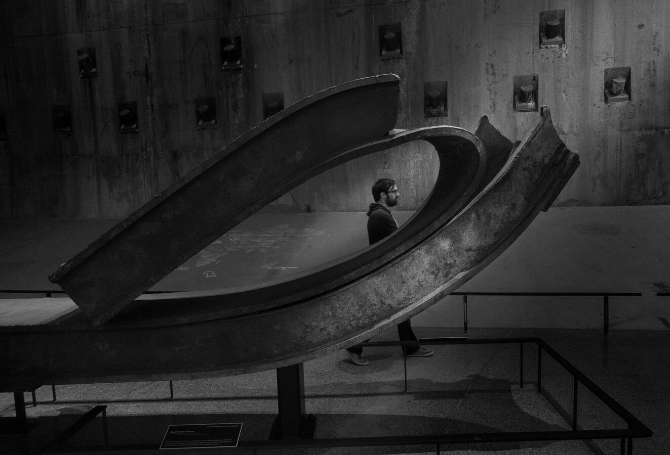 A somber black and white photograph from the 9/11 Museum. A man walks behind a massive, mangled iron girder, the scale of the wreckage framing his silhouette against concrete.