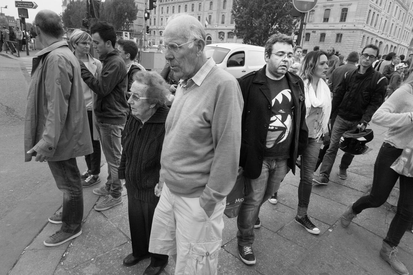A layered black and white street scene in Paris. An elderly couple pauses at a corner, while directly behind them, a man wearing a t-shirt with a scary face graphic creates a jarring contrast.