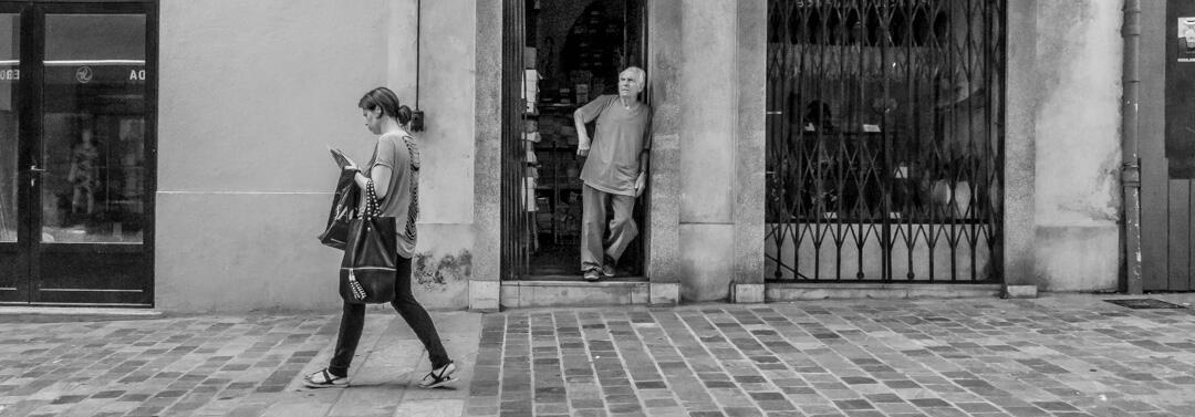 A wide black and white street scene in Bastia. An elderly shop owner leans against his doorway, watching a woman walk past while she is preoccupied with her mobile phone.