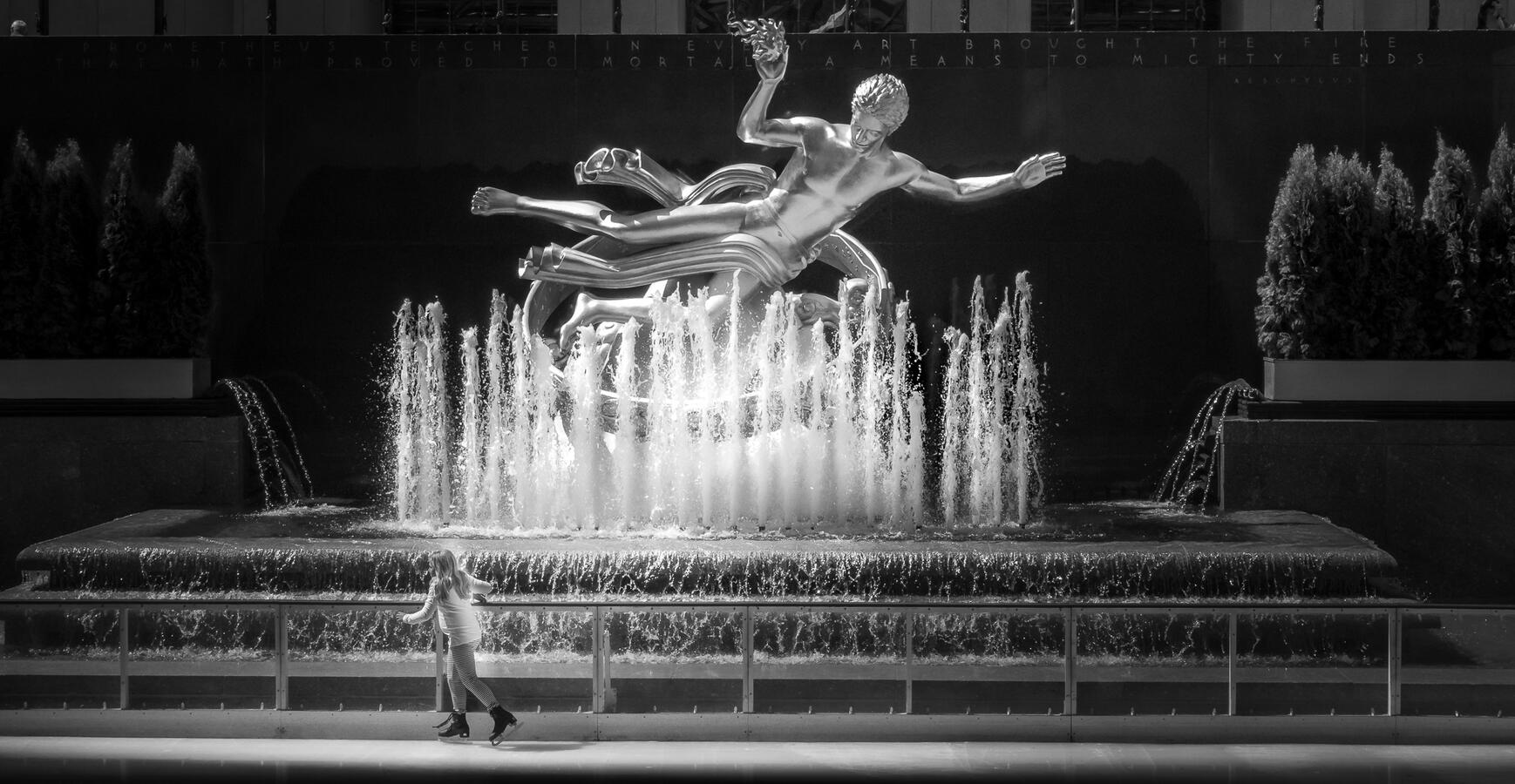 A high-contrast black and white shot at Rockefeller Center. A young girl ice skates across the bottom while the gilded Prometheus fountain sculpture looms above as if observing her.