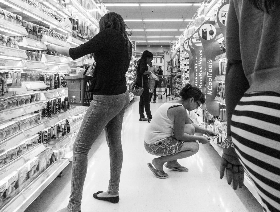 A wide-angle black and white street photograph in a Woodbridge Walmart. Four women are captured in a candid sequence of standing and squatting poses as they browse the makeup aisle, creating a repetitive visual rhythm.
