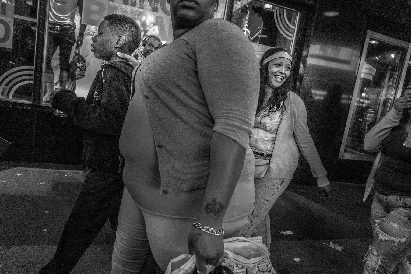 A gritty, wide-angle black and white street scene in NYC. A woman holds a young boy by the neck as they navigate a crowded sidewalk, captured with a dynamic, close-up perspective.