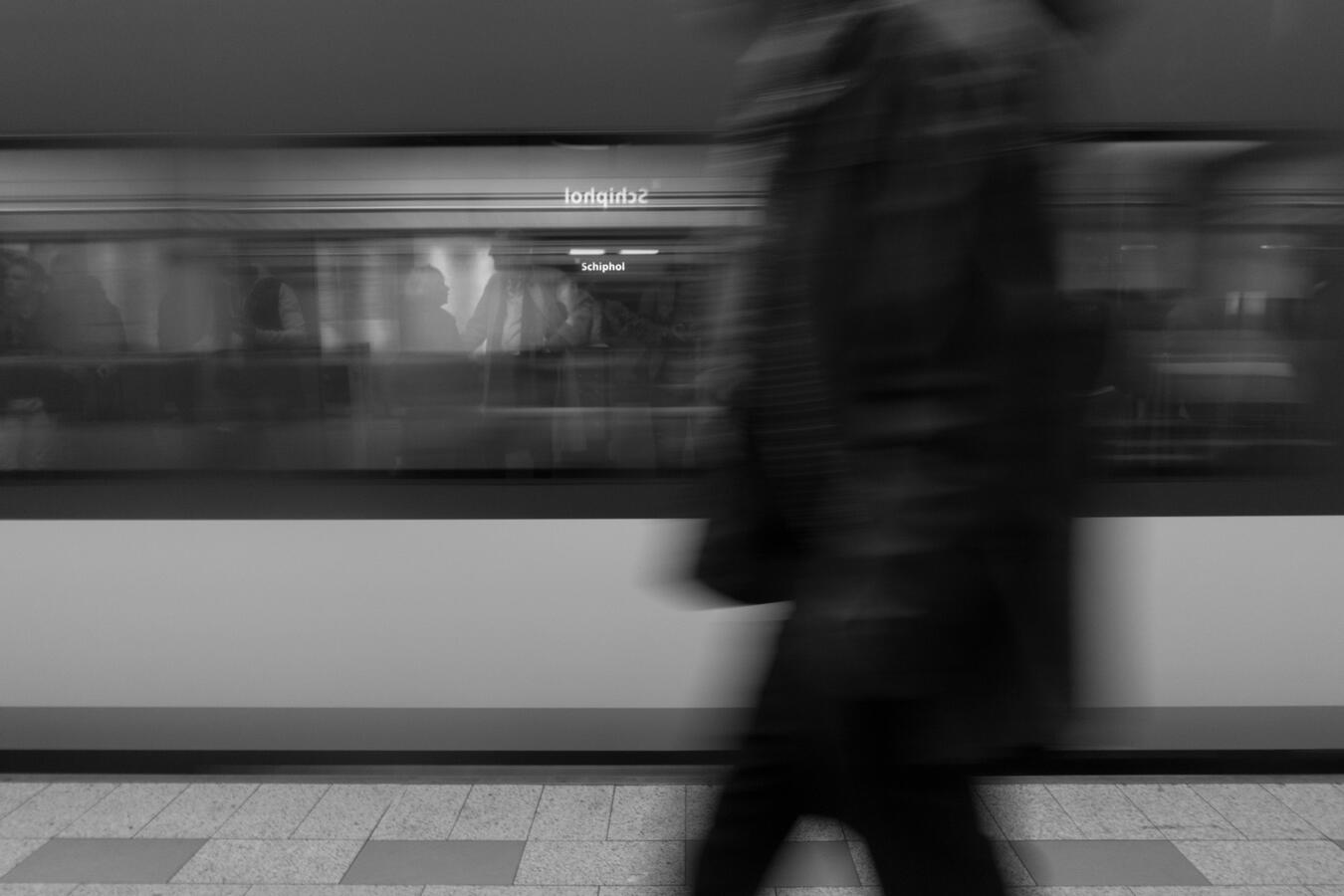 A cinematic black and white long exposure at Schiphol station. A blurred figure in the foreground passes a train window where a couple is seen talking in sharp silhouette.