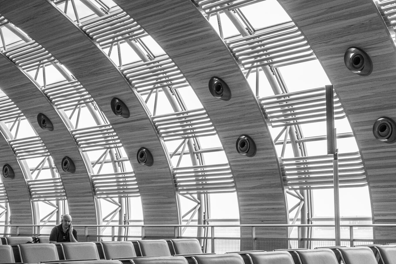 A black and white architectural study of the CDG Airport terminal. A lone man sits in the lower left, dwarfed by the massive, rhythmic wooden arches and structural ribs of the ceiling.