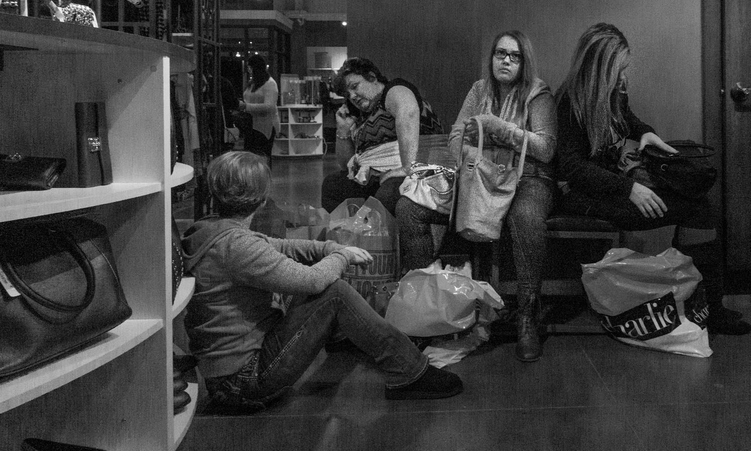 A gritty black and white street photograph in Woodbridge. Four women find a spot to sit and rest on the floor and benches during an all-night Christmas shopping event.