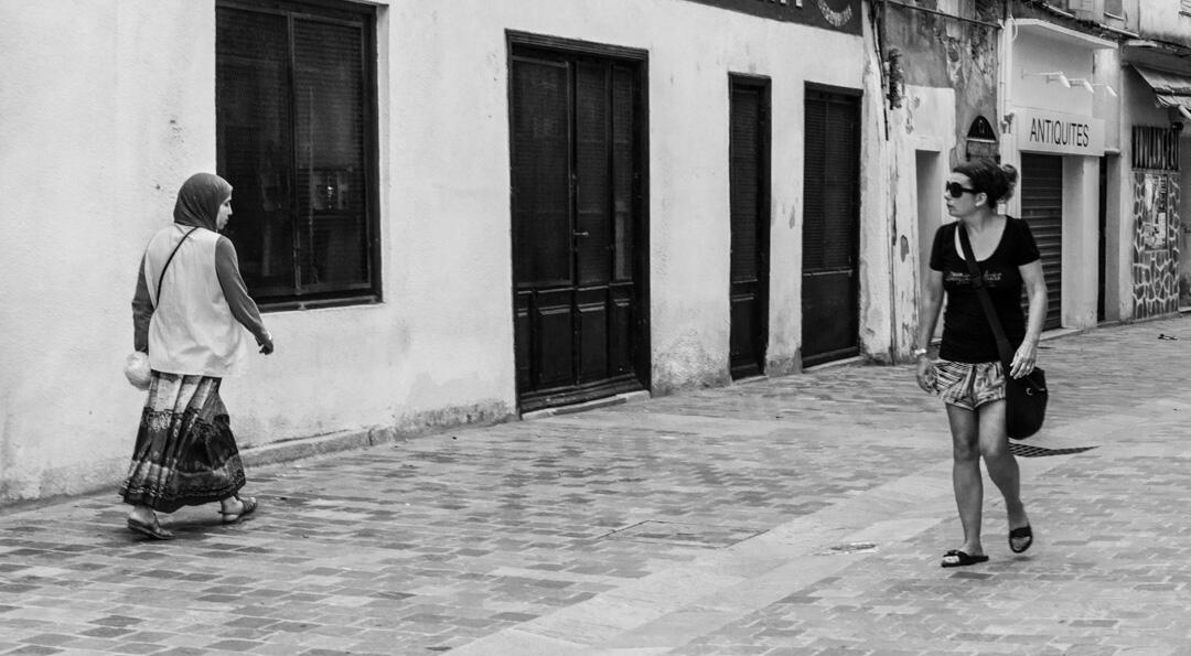 A black and white street observation in Bastia. Two women walk past each other; one in traditional Middle Eastern attire and the other in modern shorts, creating a shared moment of visual contrast.