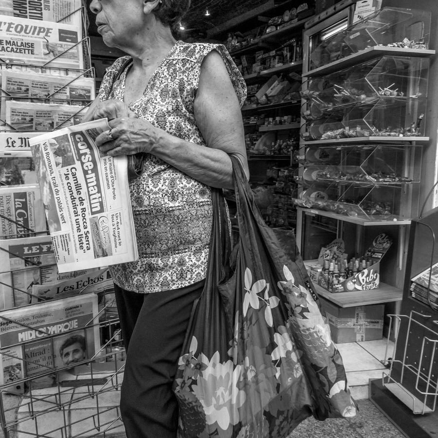 A candid black and white shot in Bastia. A woman exits a shop holding a copy of the Corse-Matin newspaper, her floral shopping bag adding a complex pattern to the busy shop interior.