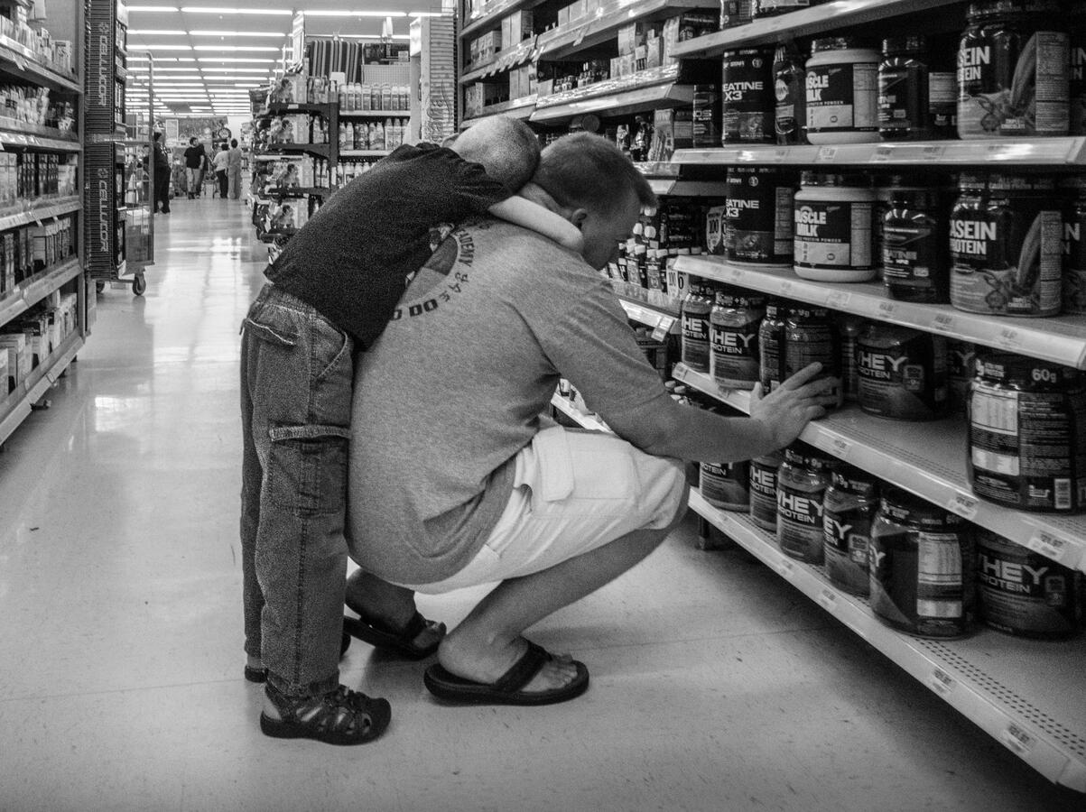A candid black and white moment in a Woodbridge Walmart. A young boy with a crew cut leans over his father's shoulder as the man squats to examine protein powder on a lower shelf.
