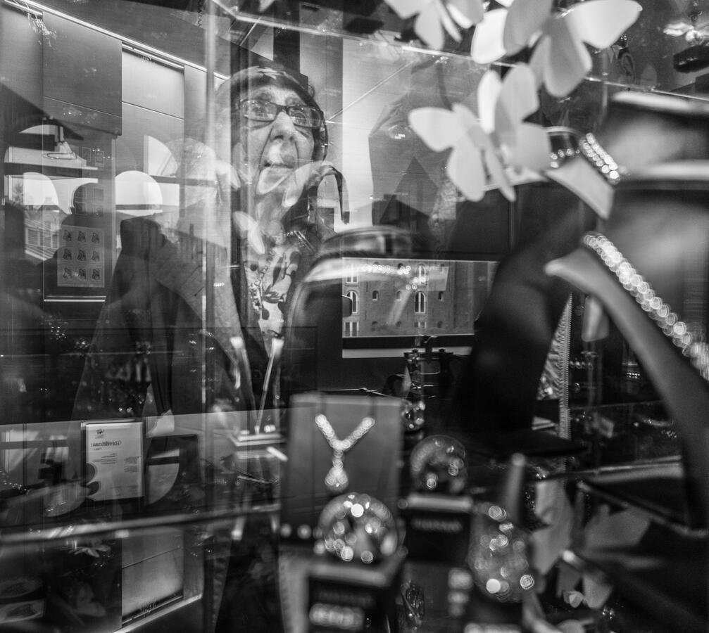 A complex black and white street photograph in Amsterdam. An elderly woman’s face is reflected in a jewelry store window as she peers intently through the glass at a display.