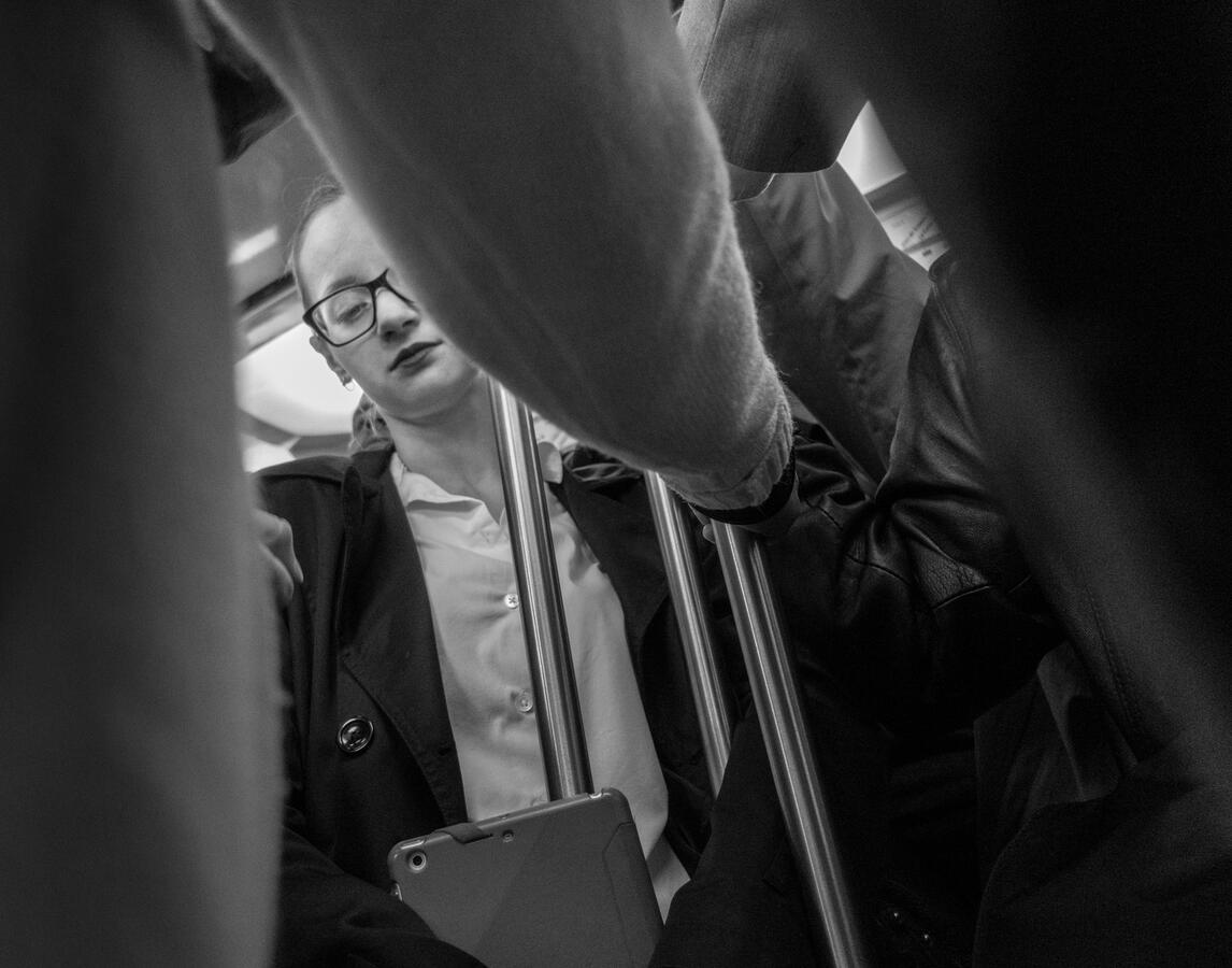 A gritty, close-up black and white street photo inside the Paris Metro. A young woman with glasses stares off into space, framed by the dark shapes of other commuters.